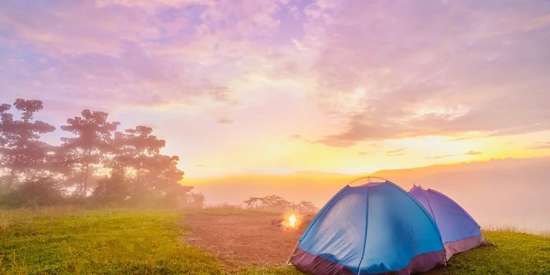 Two tents on a grassy hill at sunrise with a distant forest and morning mist
