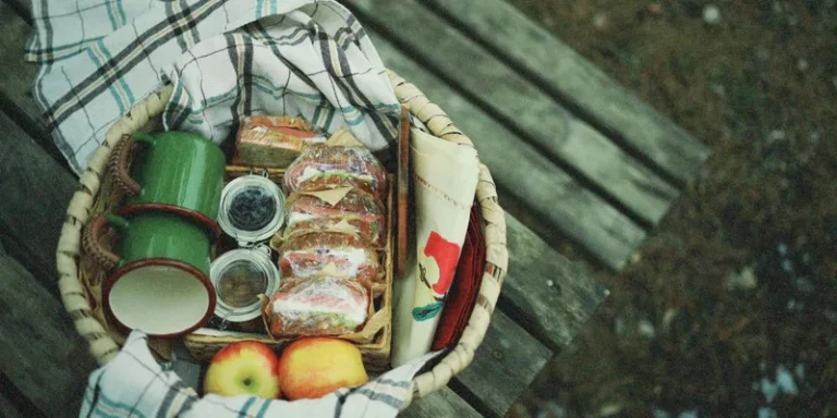 A cozy picnic basket with sandwiches, apples, and cups on a wooden bench outdoors
