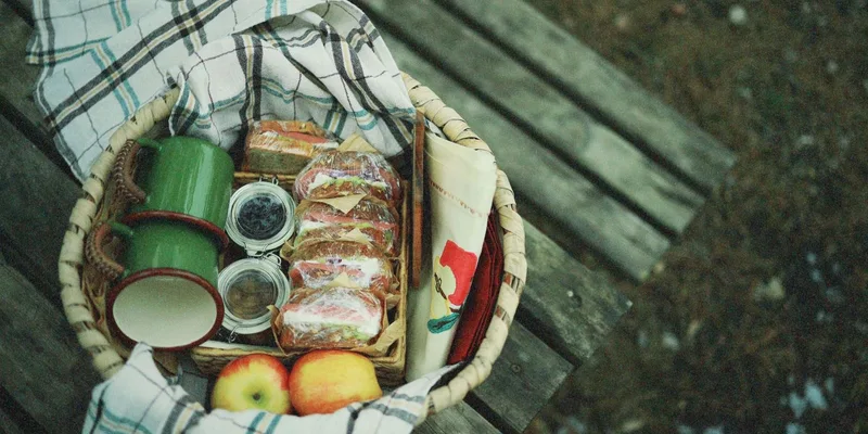 A cozy picnic basket with sandwiches, apples, and cups on a wooden bench outdoors