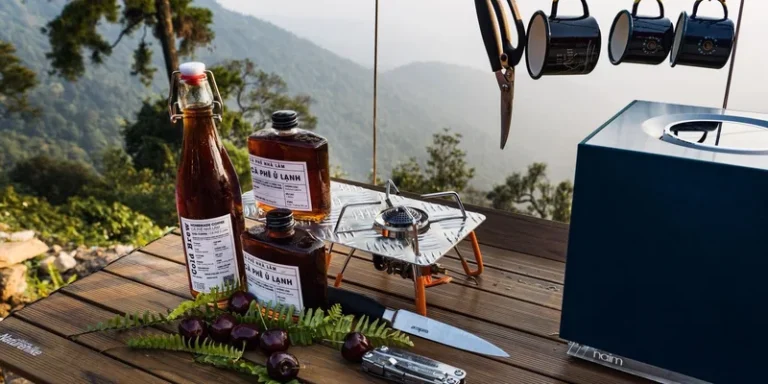 Camping coffee setup with bottles, mugs, and tools on a wooden table against a mountain backdrop in Vĩnh Phúc, Vietnam