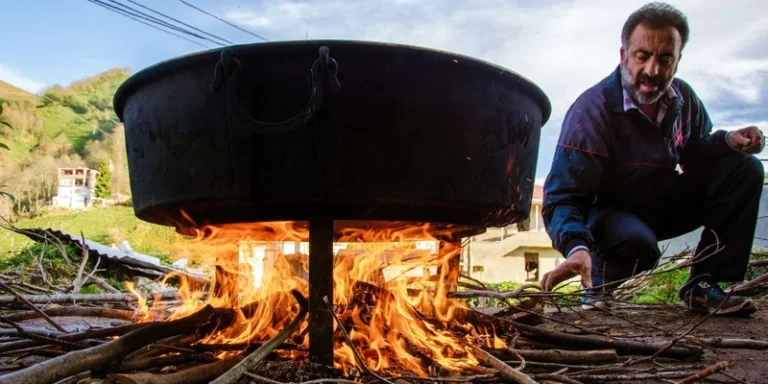A man tends to an outdoor fire cooking in a large cauldron amidst a natural setting