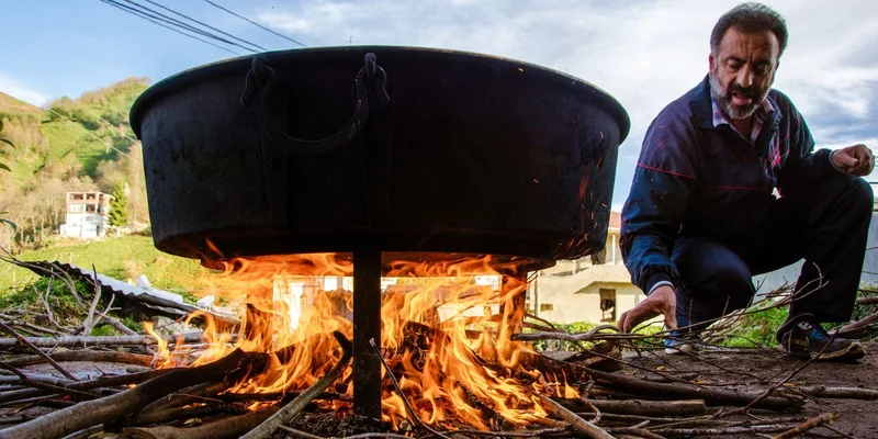 A man tends to an outdoor fire cooking in a large cauldron amidst a natural setting