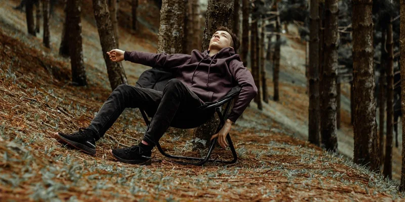 A man in a hoodie rests on a folding chair in a serene forest setting, enjoying leisure time