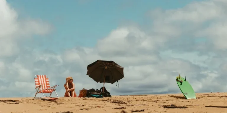 Person enjoying a sunny beach day with a chair, umbrella, and surfboard