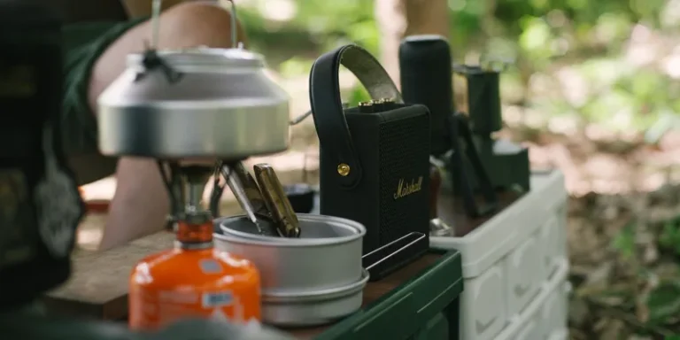 Outdoor camping gear including a gas stove and kettle on a table in a forest setting