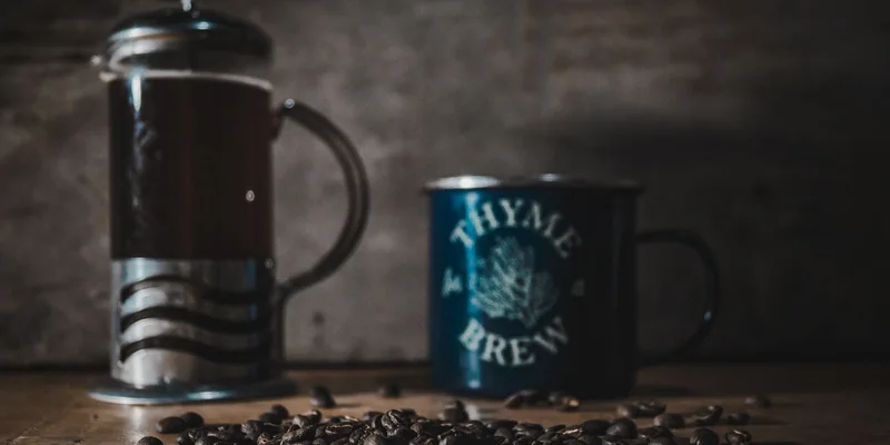 A cozy scene featuring a French press, a mug labeled 'Thyme for Brew', and coffee beans on a wooden table