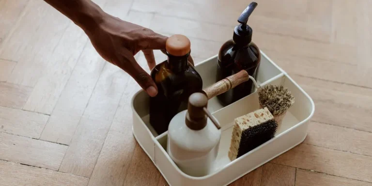 A hand reaching for a cleaning set with bottles and brushes on a wooden floor