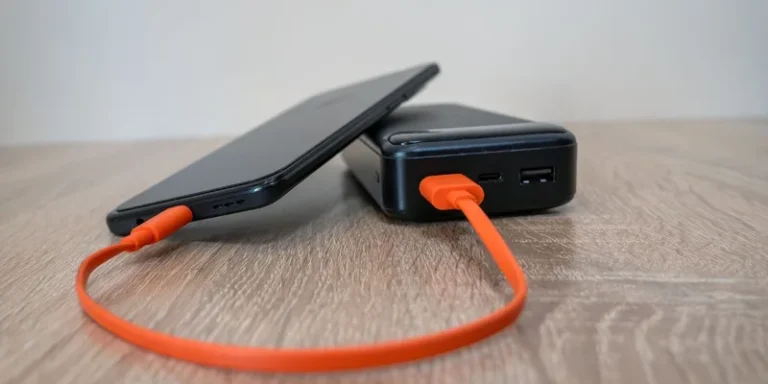 A smartphone charging from a power bank with an orange cable on a wooden table