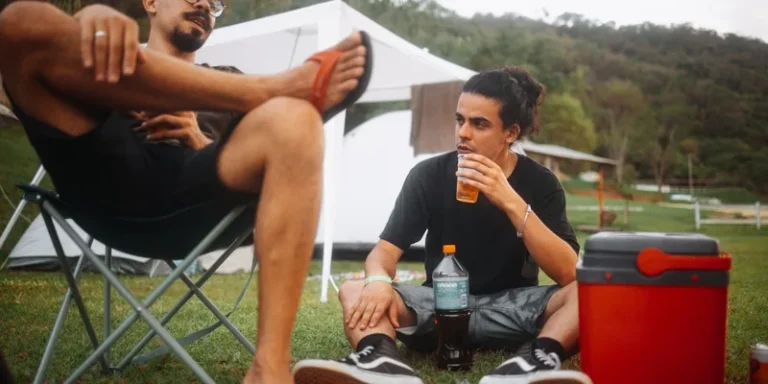 Two young men enjoying a picnic outdoors, sitting on the grass with a cooler and drinks