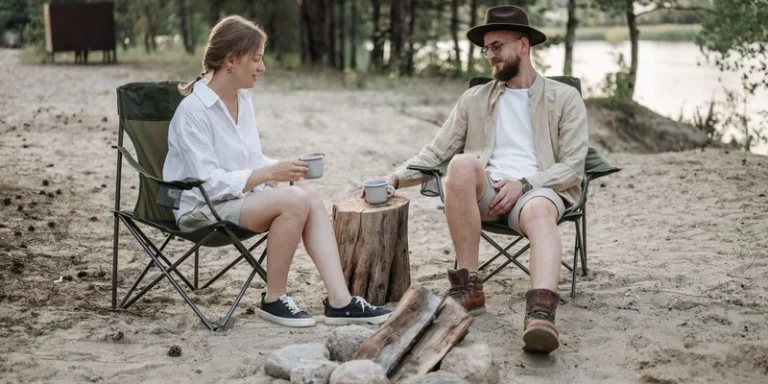 A couple relaxes with coffee by a campfire surrounded by nature