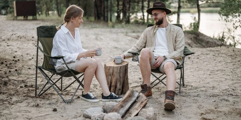 A couple relaxes with coffee by a campfire surrounded by nature