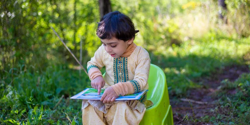 A young boy wearing traditional clothes reads a book outdoors on a green chair
