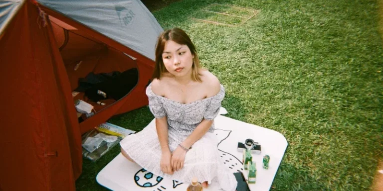A woman in a summer dress sits on a mat outside a tent, surrounded by summer scenery