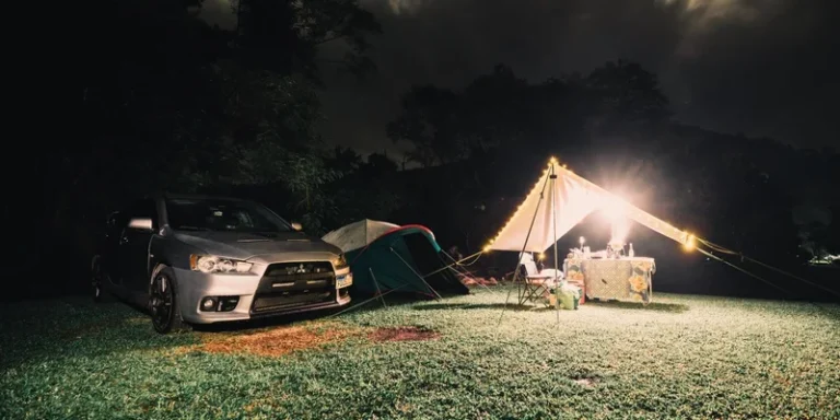Serene camping site at night featuring illuminated tents, car, and lush grass under a cloudy sky