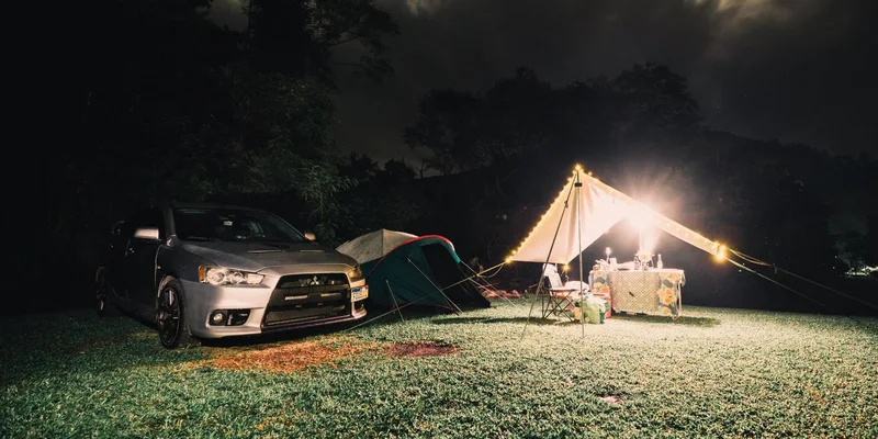 Serene camping site at night featuring illuminated tents, car, and lush grass under a cloudy sky